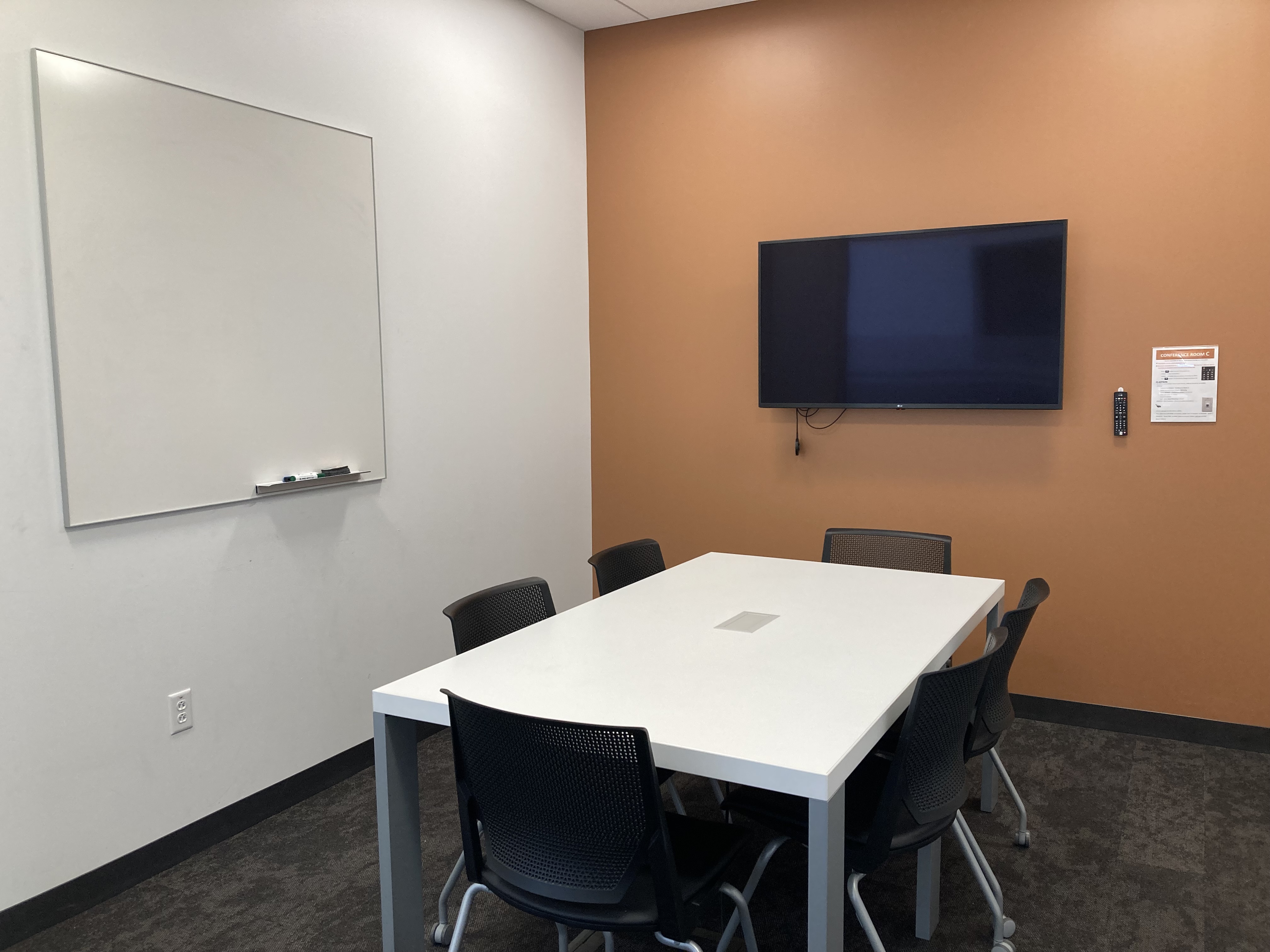 mage of conference room with white table, six chairs, white board on wall and mounted television on back orange wall.