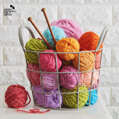 Photo of a wire basket full of different colored yarn and a set of knitting needles sitting on a table with a white brick background