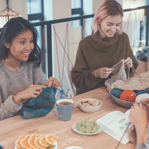 Photo graph of 2 women knitting with the hands of a 3rd woman showing across the table.