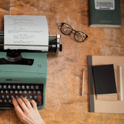Image of a desk with a typewriter being used by a human hand, to the right of that is a notebook with pencils and a pair of glasses sitting slight above the notebook