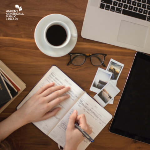 Photo taken from above of someone at a desk writing in a notebook with coffee, a laptop glasses and photos