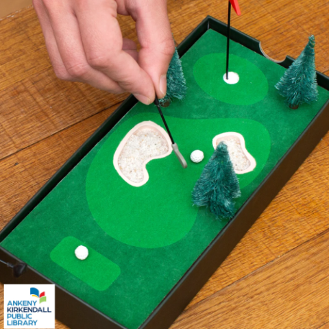 a hand pushing a miniature golf club to hit a miniature ball into a hole on the green of a miniature golf course. The Ankeny Kirkendall Public Library logo is in the bottom left corner.