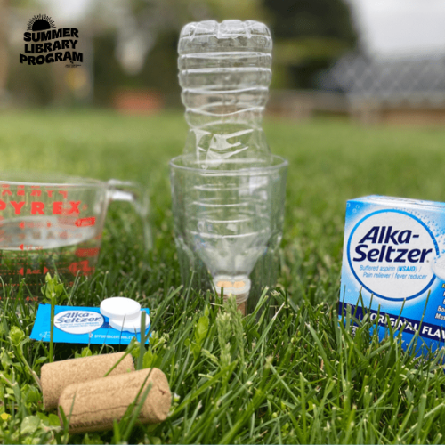 a water bottle, alka-seltzer, a measuring cup, and cork on grass
